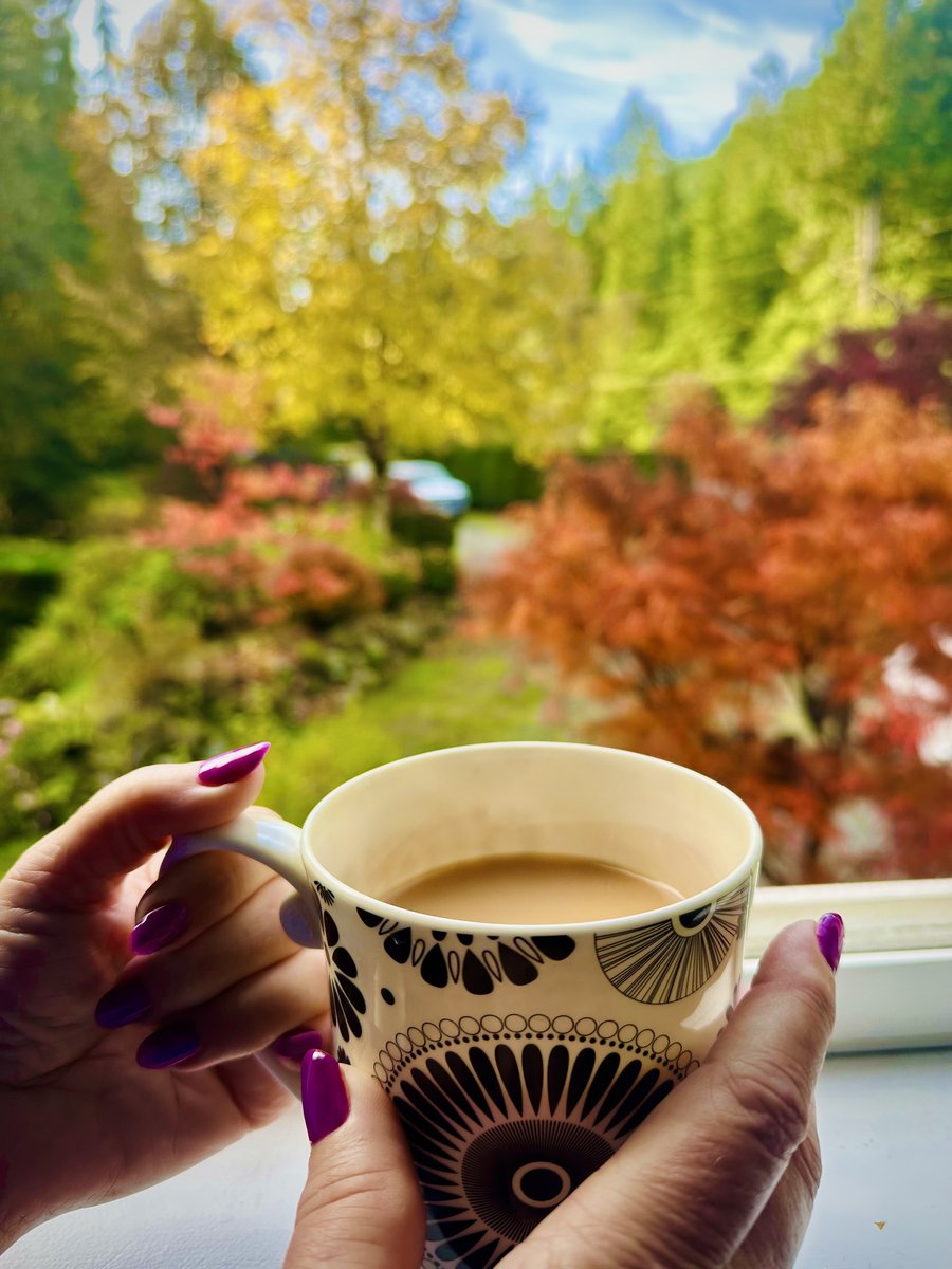 Two hands holding a patterned mug of milky tea in front of an autumn garden